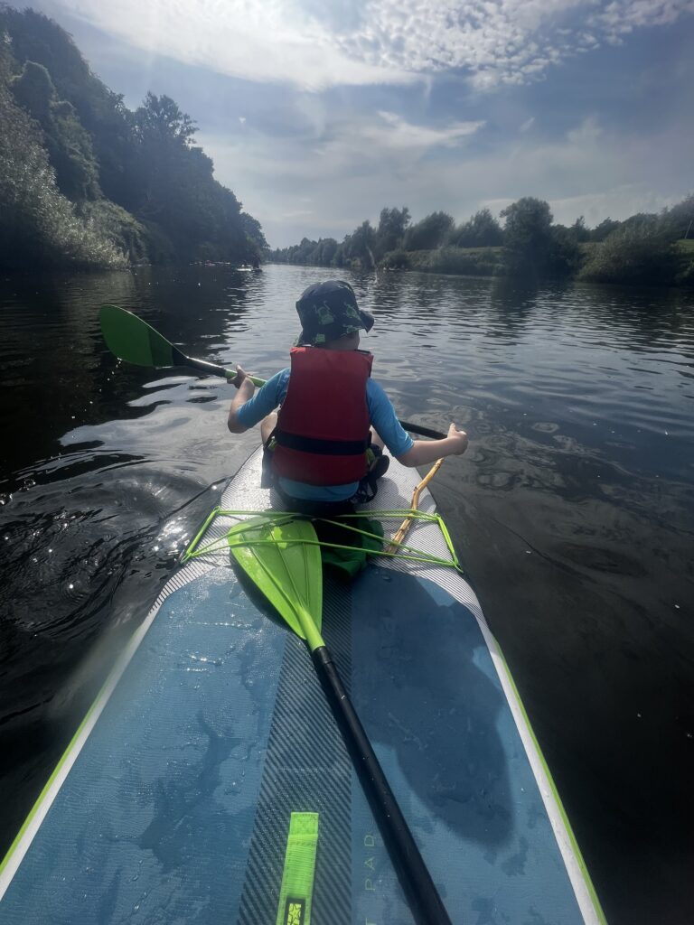 A child sat on the front of a stand-up paddleboard with a paddle on a river.