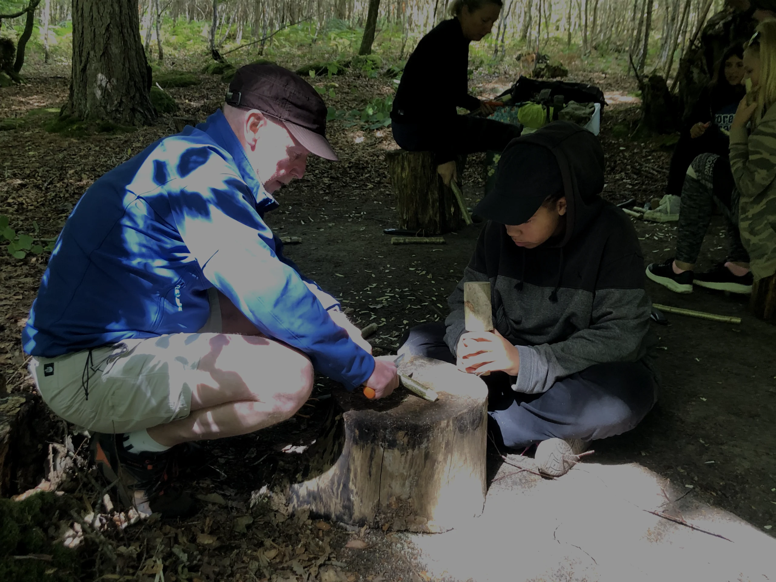 A man in a blue jacket demonstrating whittling a stick with a knife to a student.
