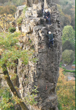 Two rock climbers ascend a narrow, jagged stone spire rising from a forested hillside, with trees and autumn-colored foliage visible in the background below.