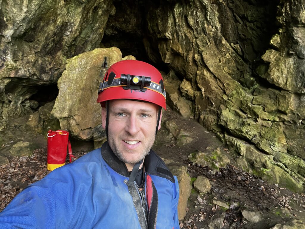A photo of a Ed Perry, the owner of forest adventure, in blue overalls, a red helmet and torch in front of a cave entrance.