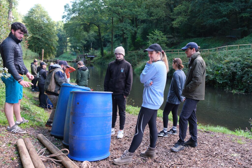 A group of people in casual outdoor clothing standing next to a riverbank with two large blue barrels in the foreground, surrounded by trees.
