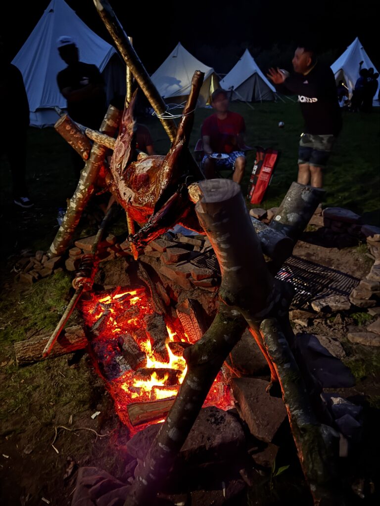 A whole animal is slow-roasted over an open campfire at night, suspended on wooden poles above glowing embers, with people and canvas tents gathered in the background.