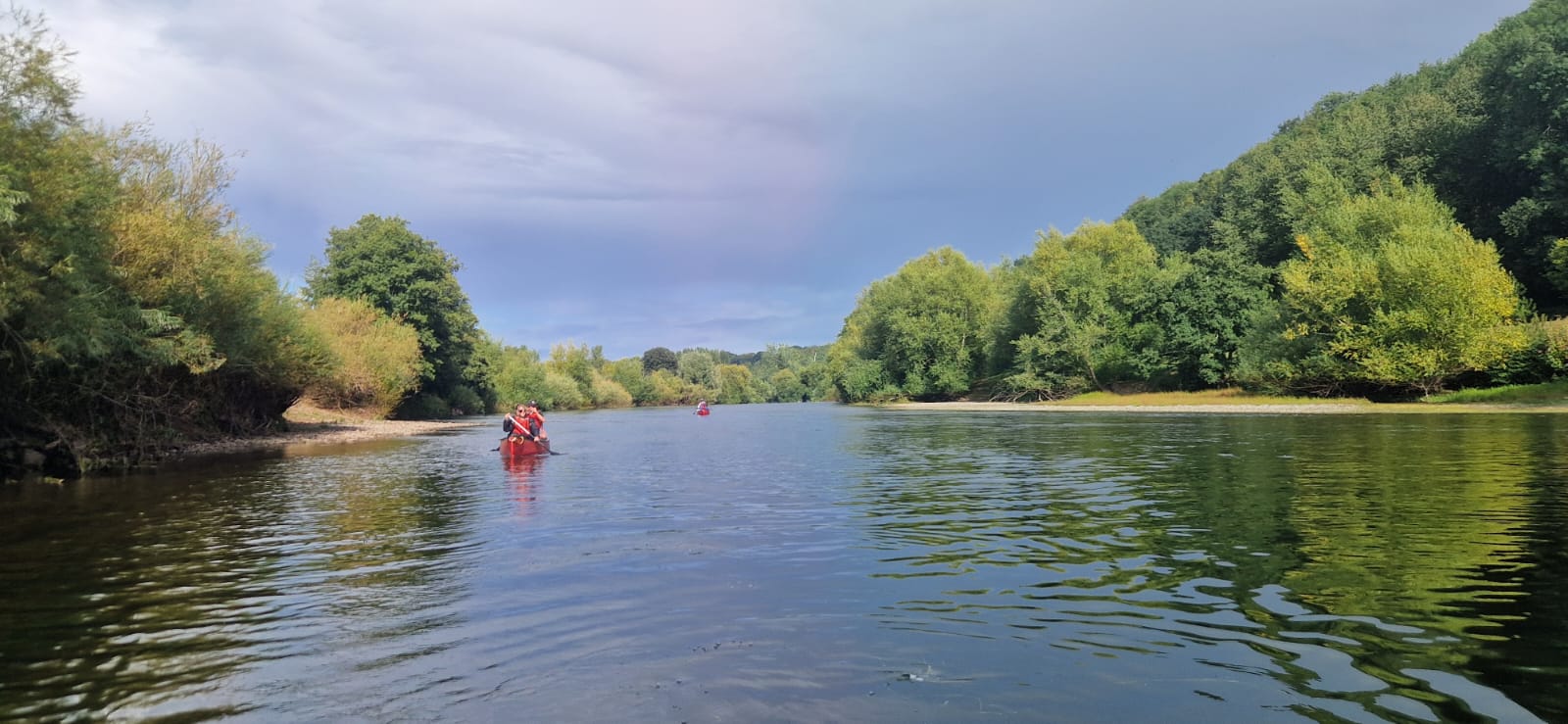 Canoeists paddle along a wide, calm river bordered by dense green woodland under a lightly clouded sky.