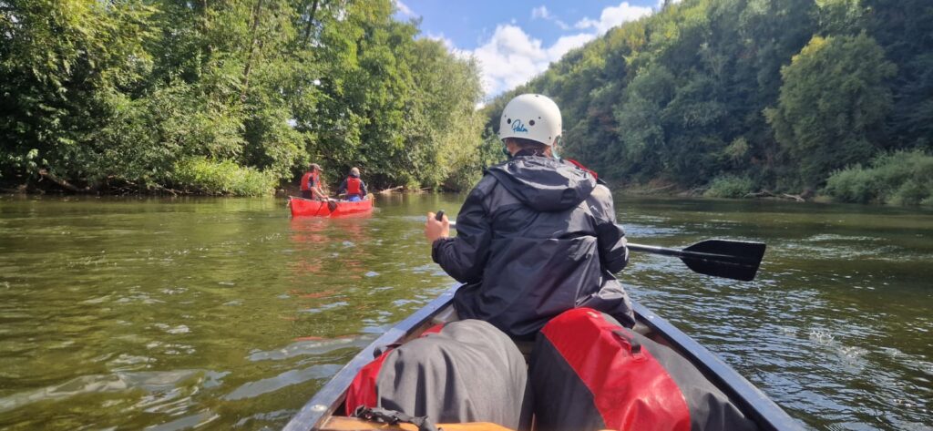 Person paddling a canoe along a calm river with a group ahead surrounded by trees