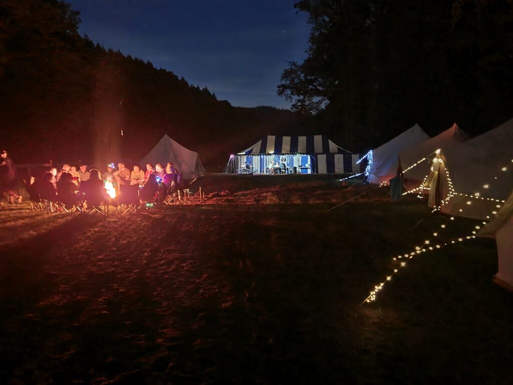 People gather around a campfire at night in a riverside campsite with canvas tents, string lights, and a large striped marquee tent glowing in the background.