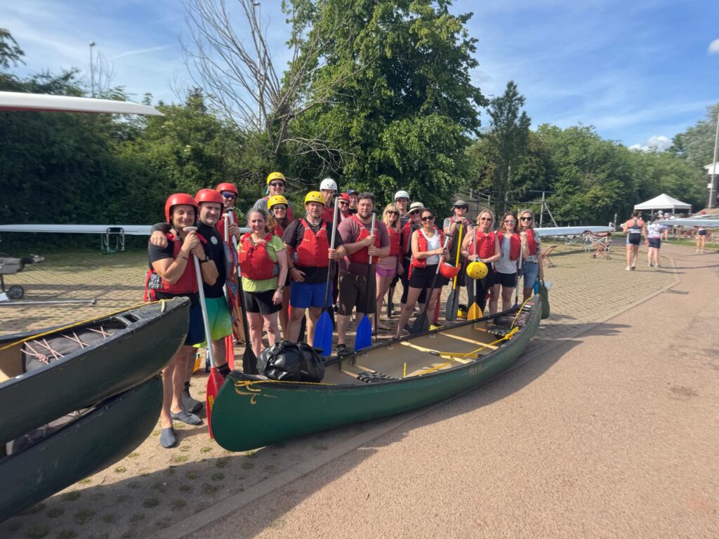 A large group of adults wearing life jackets and helmets pose with canoes and paddles beside a riverside launch area under clear blue skies.